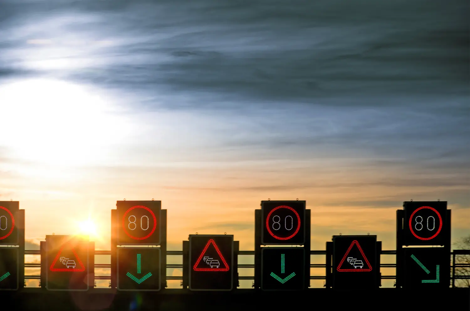 Logos Ingenieurplanungs GmbH – Das Bild zeigt eine Reihe elektronischer Verkehrsleittafeln über einer Autobahn, die wahrscheinlich Fahrstreifensperrungen oder Geschwindigkeitsbegrenzungen zur Verkehrsregelung anzeigen.